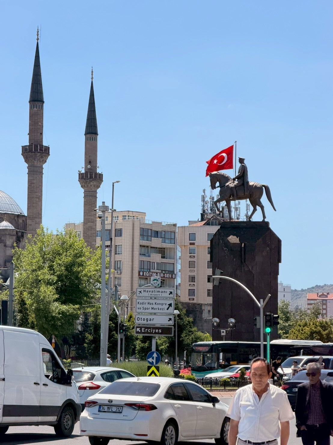 トルコ国旗を掲げた騎馬像と、街を行き交う車。背景に2本のミナレットが見える。
